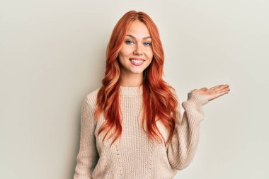 Genç, güzel, kızıl saçlı, sıradan bir kazak giyen neşeli bir şekilde gülümseyen ve avuç içi kameraya bakan bir kadın.. 