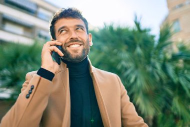 Zarif bir ceket giyen yakışıklı bir iş adamı akıllı telefon kullanarak dışarıda mutlu bir şekilde gülümsüyor.