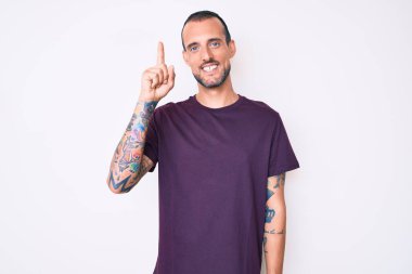 Dövmeli genç ve yakışıklı bir adam günlük kıyafetler giyiyor. Bir numarayı gösteriyor ve işaret ediyor. Kendinden emin ve mutlu bir şekilde gülümsüyor.. 