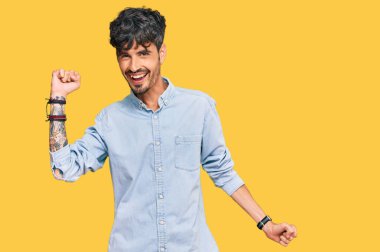 Genç İspanyol adam gündelik kıyafetler giyip dans ediyor mutlu ve neşeli, gülümsüyor rahat ve kendinden emin bir şekilde müzik dinliyor. 