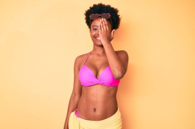 Genç Afro-Amerikan kadın bikini giyiyor. Bir gözünü eliyle kapatıyor. Yüzünde kendine güvenen bir gülümseme ve sürpriz bir duygu var.. 