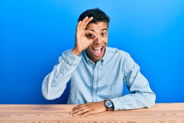 Genç, yakışıklı, Afro-Amerikalı, günlük giysiler giymiş. Masada oturuyor. El hareketleriyle gülümsüyor ve mutlu yüzüyle parmaklarının arasından bakıyor.. 