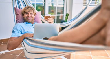 Evinin terasındaki orta yaşlı yakışıklı adam hamakta uzanıp dizüstü bilgisayarla çalışıyor ve taze bira içiyor.