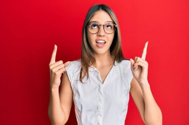 Genç esmer kadın gündelik giysiler ve gözlük takıyor. Şaşırmış ve şaşırmış bir şekilde yukarı bakıyor, parmaklarıyla işaret ediyor ve kollarını kaldırıyor.. 