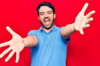 Gündelik giysiler giymiş genç İspanyol adam kameraya bakıp sarılmak için kollarını açmış gülümsüyor. Mutluluğu kucaklayan neşeli bir ifade.. 