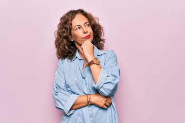 Orta yaşlı güzel bir kadın pembe arka planda dikilmiş sıradan bir kot gömlek giyiyor ve elleri çenesinde soru soruyor, düşünceli bir yüz ifadesi var. Düşünceli bir yüzle gülümsüyor. Şüphe kavramı.