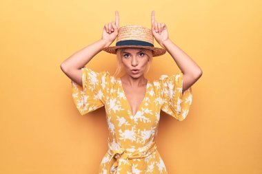 Sarışın, güzel bir kadın tatilde. Sarı arka planda şapkası ve elbisesi var. Boğanın boynuzları gibi parmağıyla komik jestler yapıyor.