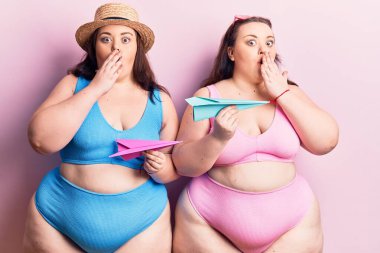 Bikini giyen genç ve büyük beden ikizler ellerinde kağıt uçakla ağızlarını kapatıyorlar, şok geçiriyorlar ve hata yapmaktan korkuyorlar. şaşırmış ifade 