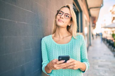 Genç sarışın kadın şehirde akıllı telefon kullanarak mutlu bir şekilde gülümsüyor..