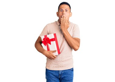 Genç, yakışıklı, Latin bir adam elinde hediyeyle ağzını eliyle kapatıyor. Şok olmuş ve hata yapmaktan korkuyor. şaşırmış ifade 