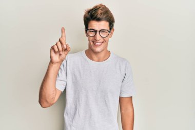 Yakışıklı beyaz adam, günlük kıyafetler ve gözlüklerle gülümsüyor bir fikir veya soruyla gülümsüyor, mutlu bir yüzle, bir numara. 