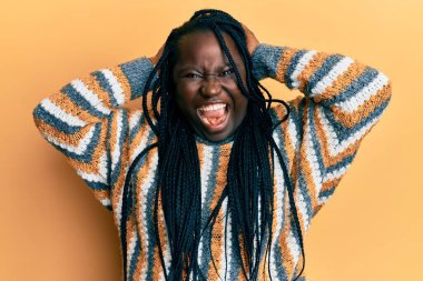 Saçları örgülü genç siyahi kadın sıradan bir kazak giyiyor çılgın ve elleri kafasında korkmuş, ağzı açık şoktan korkmuş ve şaşırmış. 