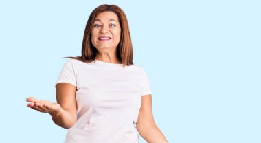 Orta yaşlı Latin kadın, günlük beyaz tişört giyiyor. Neşeli gülümsüyor, kollarını açıyor. Dostça karşılama, olumlu ve kendinden emin selamlar. 