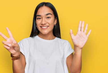 Genç Asyalı kadın, günlük beyaz tişört giyer ve yedi numaralı parmaklarıyla gülümseyerek mutlu ve kendinden emin bir şekilde onu işaret eder.. 