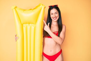 Genç, beyaz, güzel bir kadın bikini giyiyor, elinde yüzer halde gülümsüyor. 