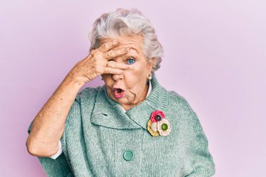 Kıdemli gri saçlı kadın, günlük giysiler giymiş. Şoka girmiş, yüzünü ve gözlerini eliyle kapatmış, parmaklarından korkuyla bakıyor. 
