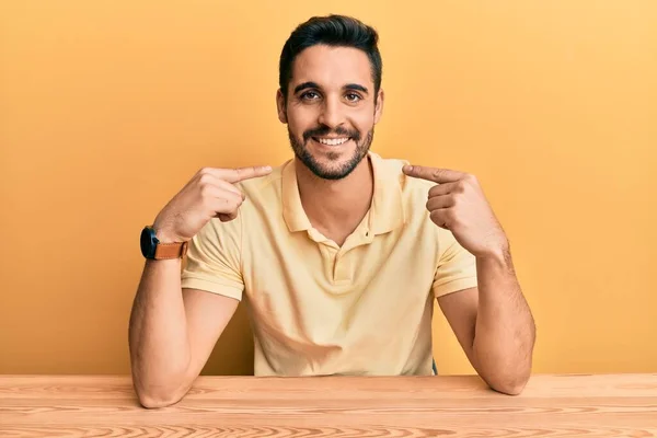 若いヒスパニック系の男性は テーブルの上に座ってカジュアルな服を着て陽気に示す笑顔と指の歯と口を指しています 歯の健康の概念 — ストック写真