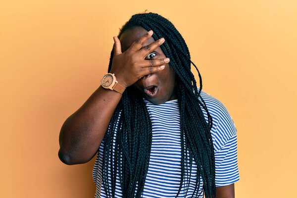Young black woman with braids wearing casual clothes peeking in shock covering face and eyes with hand, looking through fingers afraid
