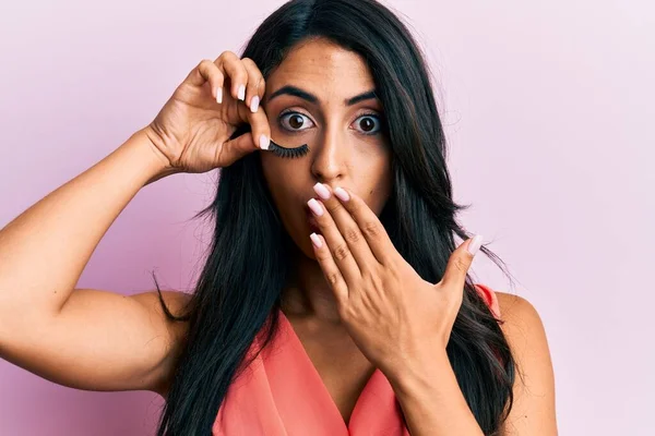 Beautiful hispanic woman holding fake lashes covering mouth with hand, shocked and afraid for mistake. surprised expression