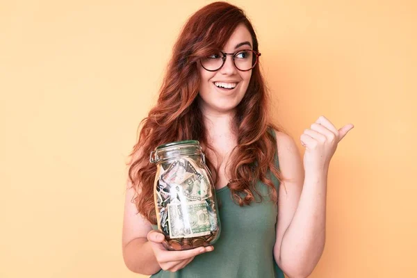 Joven Hermosa Mujer Usando Gafas Sosteniendo Frasco Con Ahorros Apuntando —  Fotos de Stock