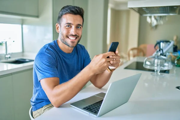 Genç Yakışıklı Adam Evde Akıllı Telefon Dizüstü Bilgisayar Kullanarak Mutlu — Stok fotoğraf