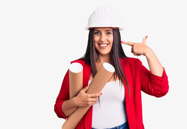 年轻美丽的黑发女子戴着硬礼帽 拿着建筑师的蓝图 指指点点着一个快乐而自豪的自我微笑的人 — 图库照片