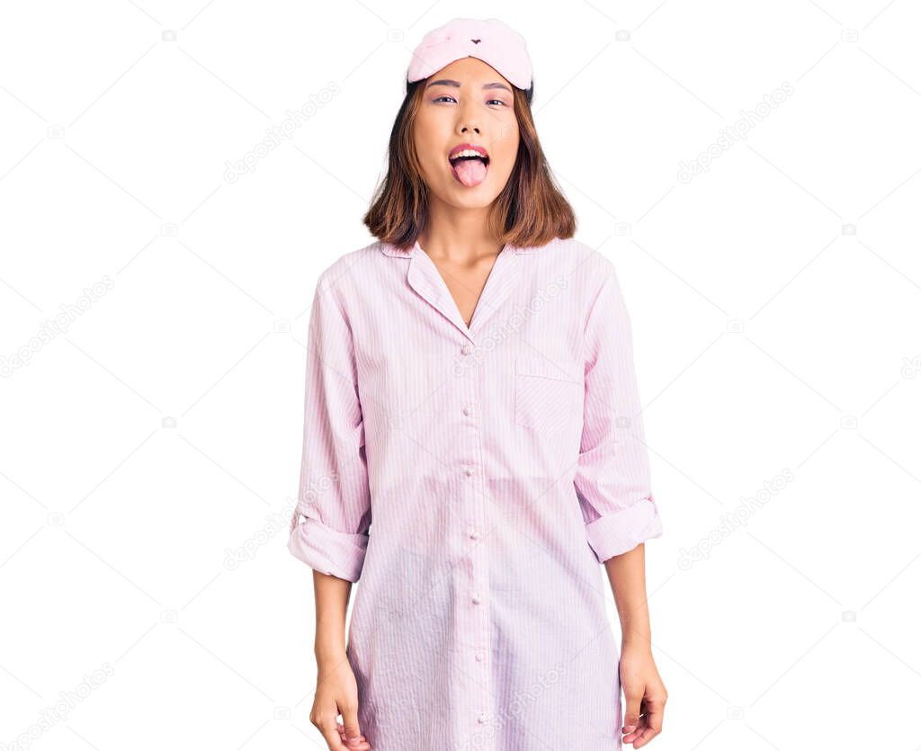 Young beautiful chinese girl wearing sleep mask and pajama sticking tongue out happy with funny expression. emotion concept. 