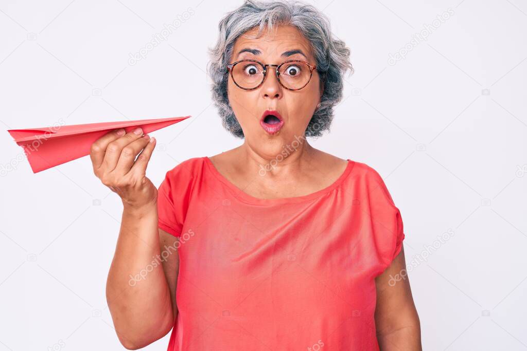 Senior hispanic grey- haired woman holding paper airplane scared and amazed with open mouth for surprise, disbelief face 