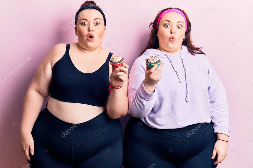 Young plus size twins wearing sportswear holding cupcake scared and amazed with open mouth for surprise, disbelief face 
