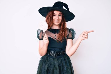 Cadı cadılar bayramı kostümü giymiş genç ve güzel bir kadın elinde lolipop tutarak gülümsüyor. 