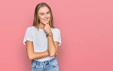 Güzel, genç, sarışın bir kadın, sıradan beyaz bir tişört giyip kameraya bakıyor. Kollarını kavuşturup gülümsüyor ve çenesini kaldırıyor. Olumlu düşünmek. 