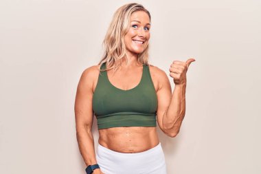 Orta yaşlı beyaz kadın spor giysisi giymiş mutlu yüzüyle gülümseyen ve baş parmağıyla yüzünü gösteren.. 