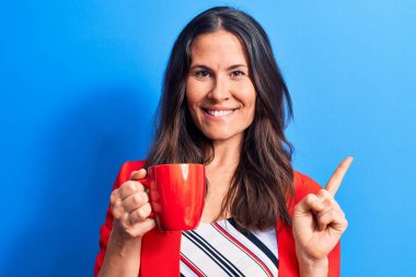 Genç ve güzel esmer iş kadını izole edilmiş mavi arka planda bir kupa kahve içerek mutlu bir şekilde elini ve parmağını kenarda gezdiriyor.