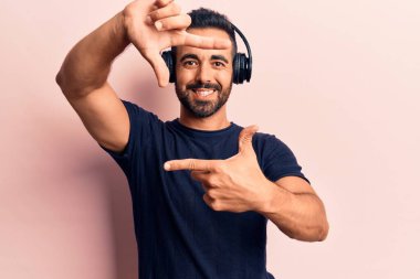Genç İspanyol adam müzik dinliyor kulaklık takıyor gülümsüyor ve elleri ve parmakları gülen yüzüyle çerçeve yapıyor. yaratıcılık ve fotoğrafçılık kavramı. 