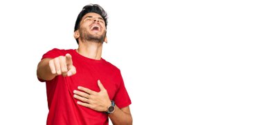 Kırmızı tişört giyen genç yakışıklı adam sana gülüyor, eli vücudunun üzerinde kamerayı işaret ediyor, utanç ifadesi. 