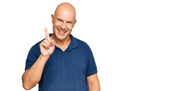 Orta yaşlı, kel, günlük giysiler giymiş bir numaralı parmağını gösterip onu gösterirken kendinden emin ve mutlu gülümsüyor.. 