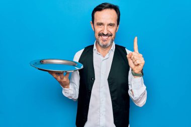 Orta yaşlı, yakışıklı garson, mavi arka planda duran tepsiyi tutarken bir fikir ya da soruyla gülümsüyor, mutlu bir yüzle işaret ediyor.