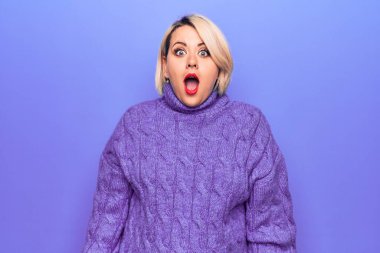 Güzel sarışın ve cüsseli bir kadın mor arka plan üzerine balıkçı yaka kazak giyiyor. Şaşırmış ve şaşırmış bir ifade, korku ve heyecanlı bir yüz ifadesi var..