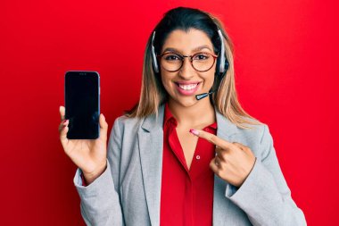 Güzel esmer kadın operatör kulaklığı takıyor. Akıllı telefon ekranını gösteriyor. Gülümsüyor. 