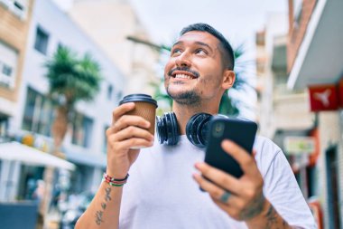 Genç İspanyol adam akıllı telefon ve kulaklık kullanıyor. Şehir caddesinde kahve içiyor.