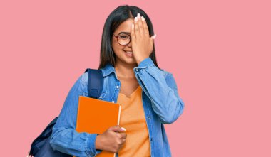 Genç Latin kız öğrenci çantası takıyor ve elinde kitaplarla bir gözünü kapatıyor, yüzünde kendine güvenen bir gülümseme ve şaşırtıcı bir duygu var.. 