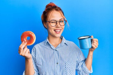 Kahvaltını yapan, çikolatalı donut ve kahve içen genç kızıl saçlı kadın kameraya seksi bir ifadeyle bakıyor, neşeli ve mutlu bir yüzle.. 
