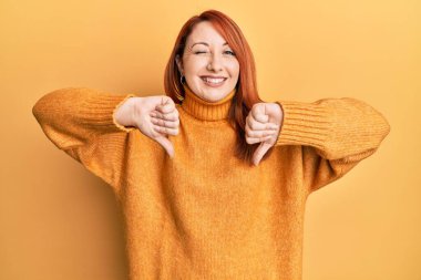 Güzel kızıl saçlı kadın negatif başparmak hareketi yapıyor. Seksi bir ifadeyle kameraya bakıyor. Neşeli ve mutlu bir yüzle.. 