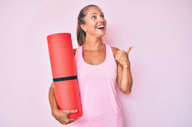 Orta yaşlı İspanyol kadın yoga minderini tutuyor başparmağını yana doğru kaldırıyor ve ağzı açık gülümsüyor. 