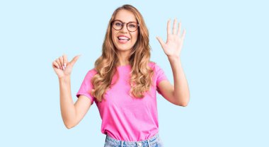 Sarı saçlı, sıradan giyinmiş, gözlüklü genç, beyaz bir kadın. Altıncı parmaklarını gösterip gösterirken kendinden emin ve mutlu bir şekilde gülümsüyor.. 