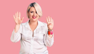 Genç, sarışın ve cüsseli bir kadın. Günlük tişört giyiyor. Dokuz numaralı parmaklarıyla işaret ediyor ve gülümsüyor.. 