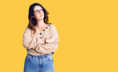 Güzel, genç, esmer, sıradan kıyafetler ve gözlük takan, kolları çapraz ve kendinden emin bir şekilde yana bakan bir kadın. 