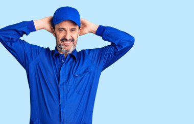 Orta yaşlı yakışıklı adam mekanik üniforma giyiyor, rahatlıyor ve esniyor, kolları ve elleri başının arkasında mutlu bir şekilde gülümsüyor. 