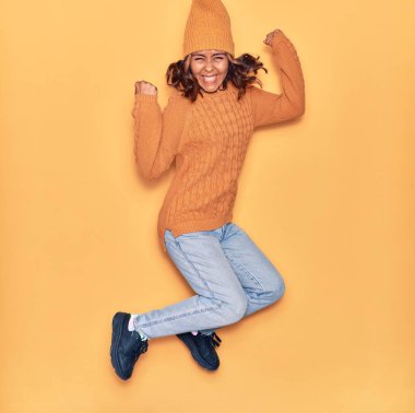 Yün şapka ve süveter giyen, mutlu gülümseyen genç, güzel Latin bir kadın. Yüzünde tebessümle zıplıyor, sarı arka planda yumruklar havada kutluyor.