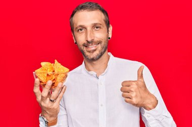 Elinde patates cipsi tutan genç yakışıklı adam mutlu ve pozitif gülümsüyor, başparmağıyla mükemmel ve onay işareti yapıyor. 
